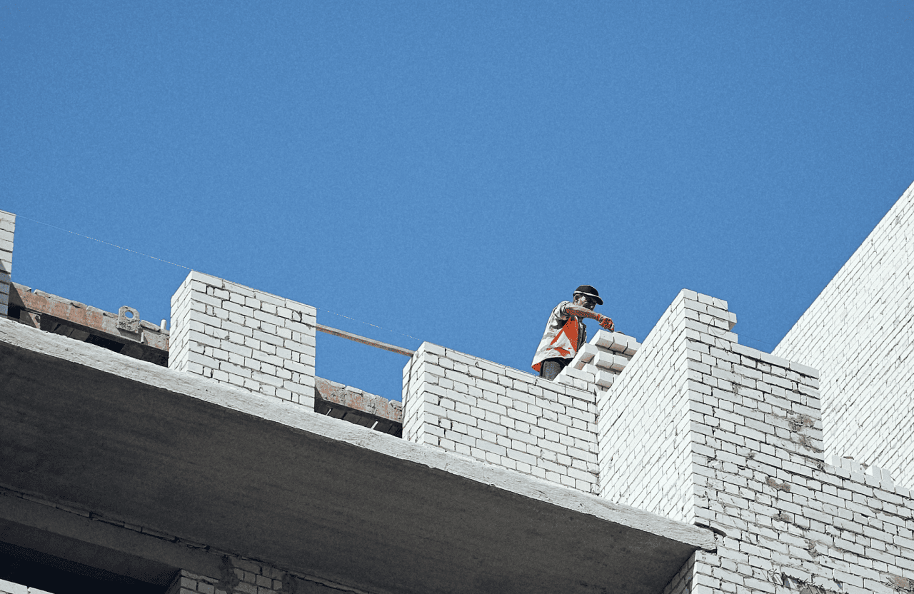 Stone Haven Developments A construction worker wearing a safety vest and helmet lays white bricks on the upper level of a building under clear blue sky. Ontario