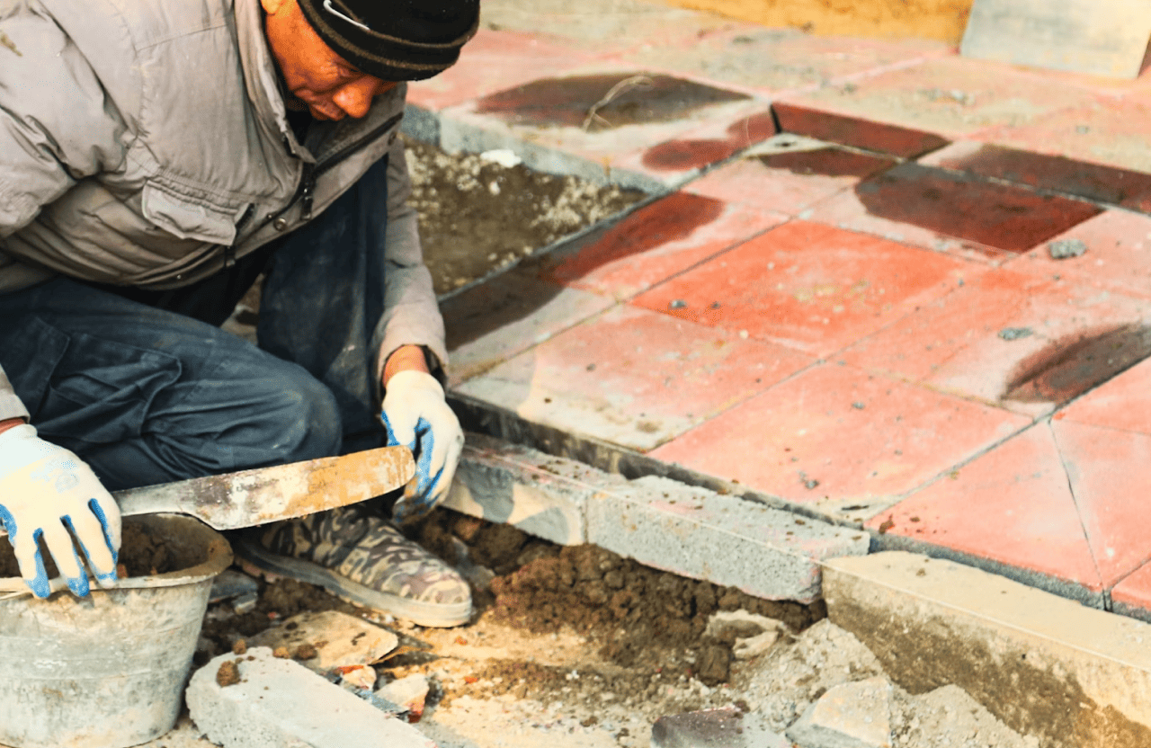 Stone Haven Developments A person wearing gloves and a jacket is using a trowel to spread mortar on the ground while laying paving stones. Ontario