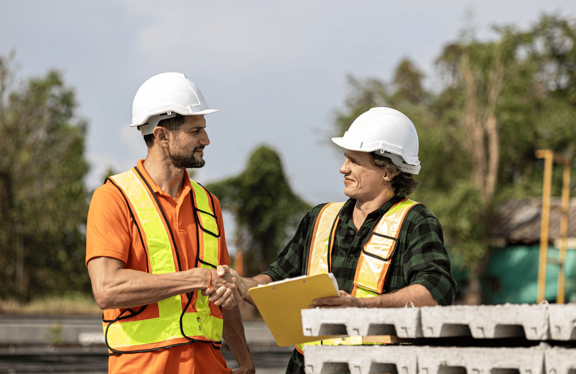 Stone Haven Developments Two construction workers wearing safety vests and helmets shake hands outdoors; one holds a yellow clipboard, with concrete blocks and trees in the background. Ontario