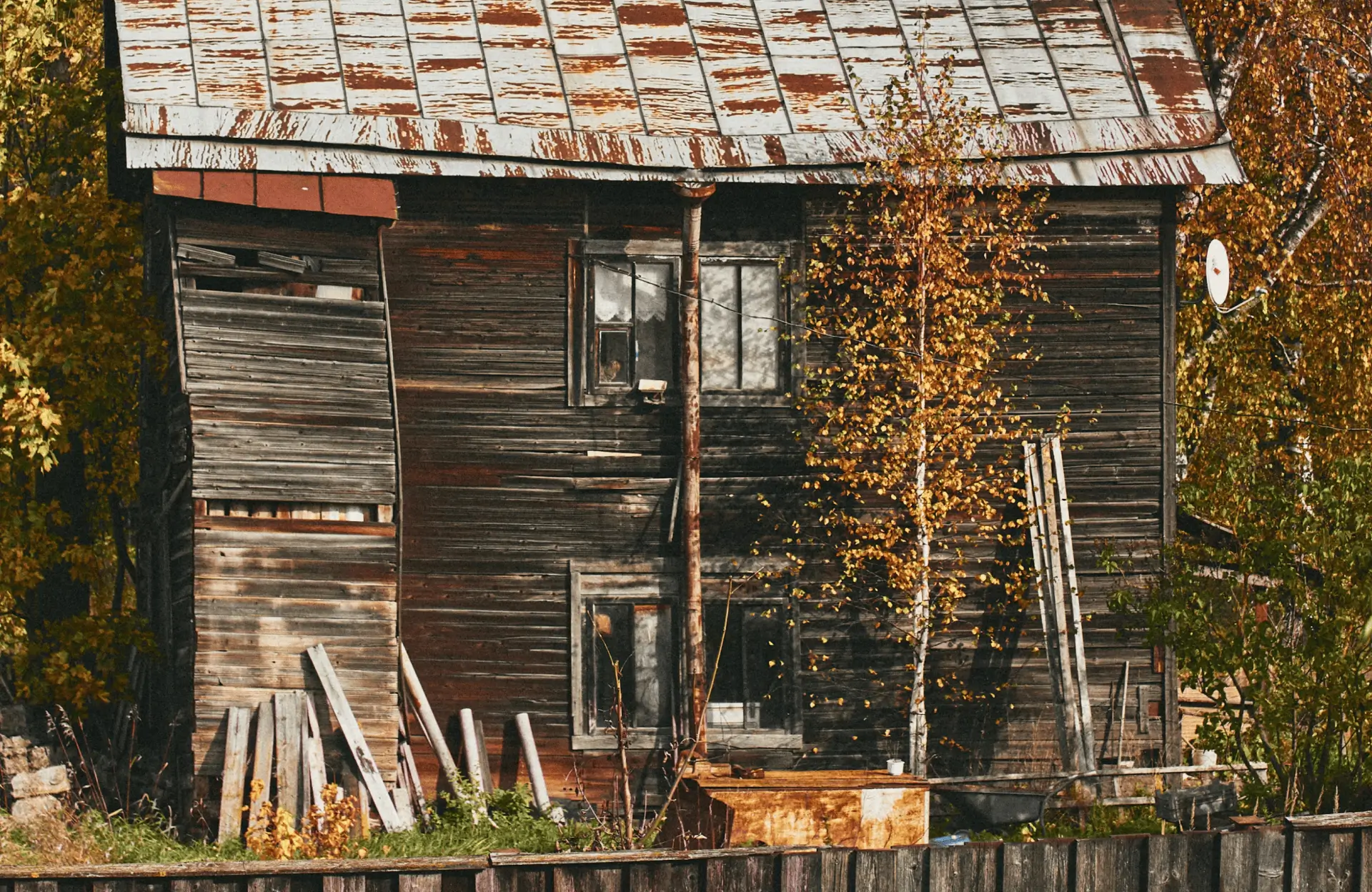 Stone Haven Developments An old, weathered wooden house with a rusty metal roof, leaning walls, and overgrown vegetation beside it, partially surrounded by a wooden fence. Ontario