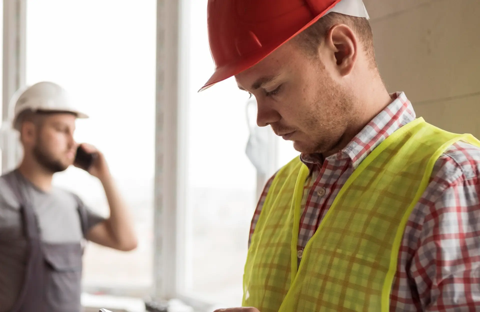 Stone Haven Developments Two construction workers indoors; one in a red hard hat and yellow vest looks down, while another in a white hard hat talks on the phone in the background. Ontario