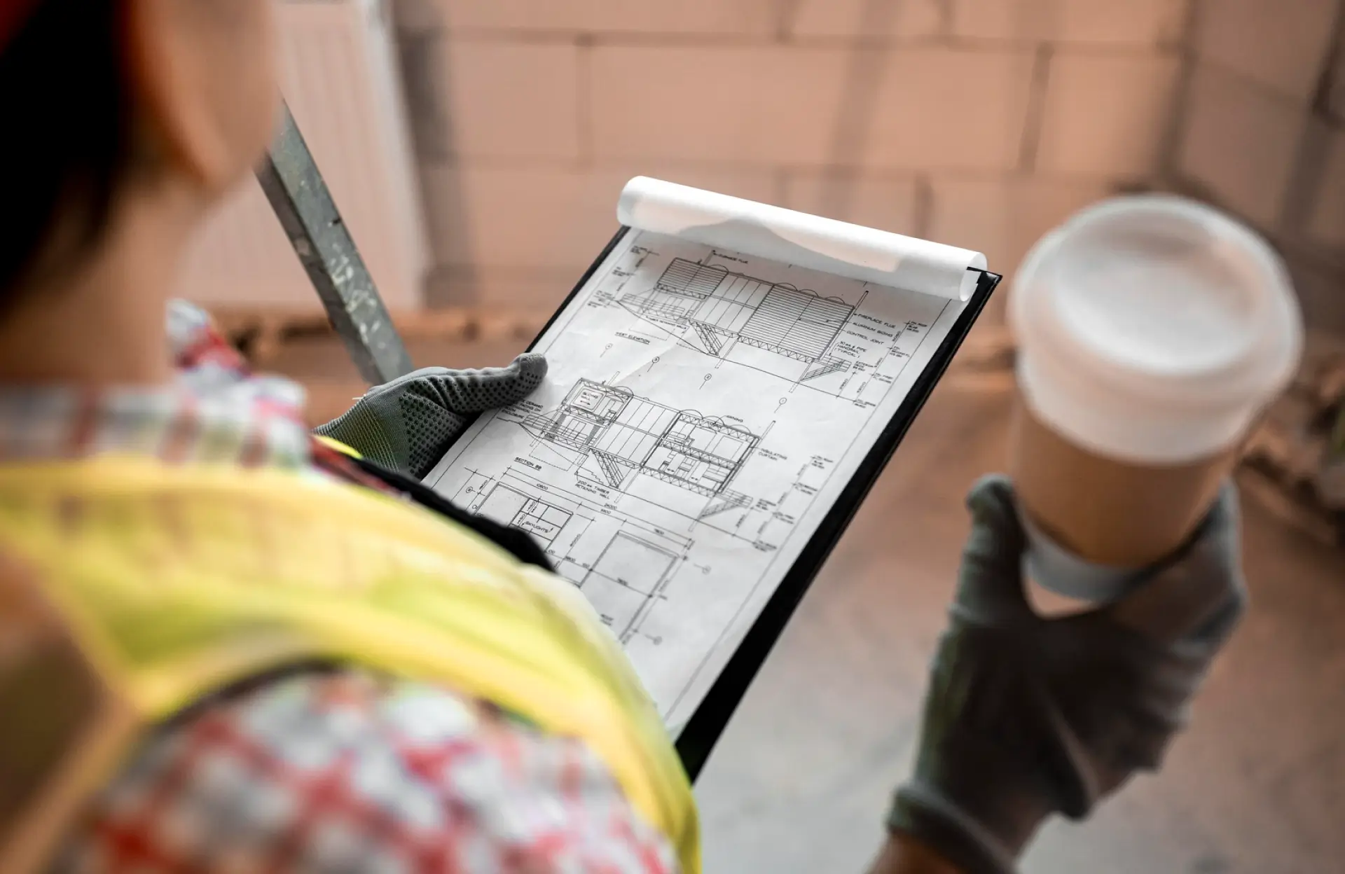 Stone Haven Developments Person in safety gear holding a clipboard with architectural blueprints in one hand and a disposable coffee cup in the other, inside a building under construction. Ontario