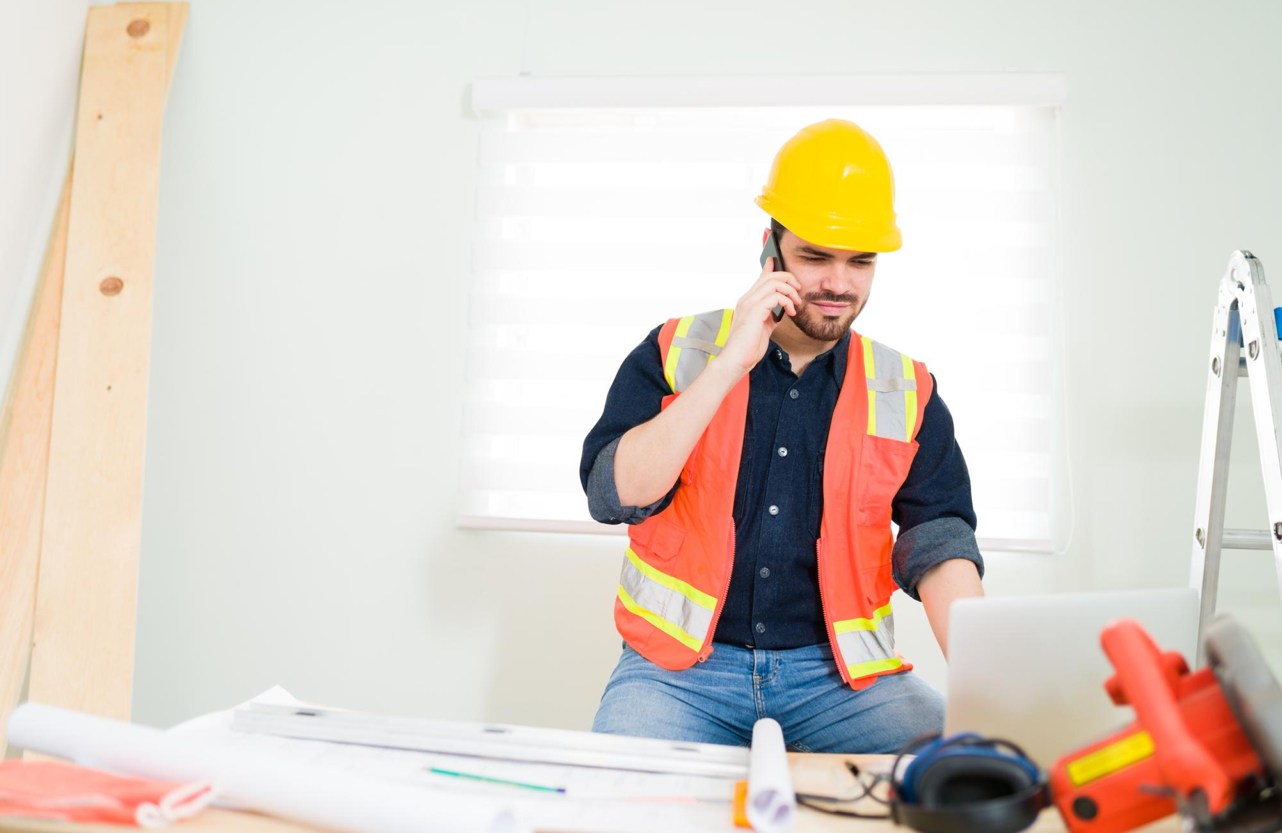 Stone Haven Developments A construction worker wearing a yellow hard hat and orange safety vest talks on the phone while using a laptop at a desk with blueprints and tools. Ontario