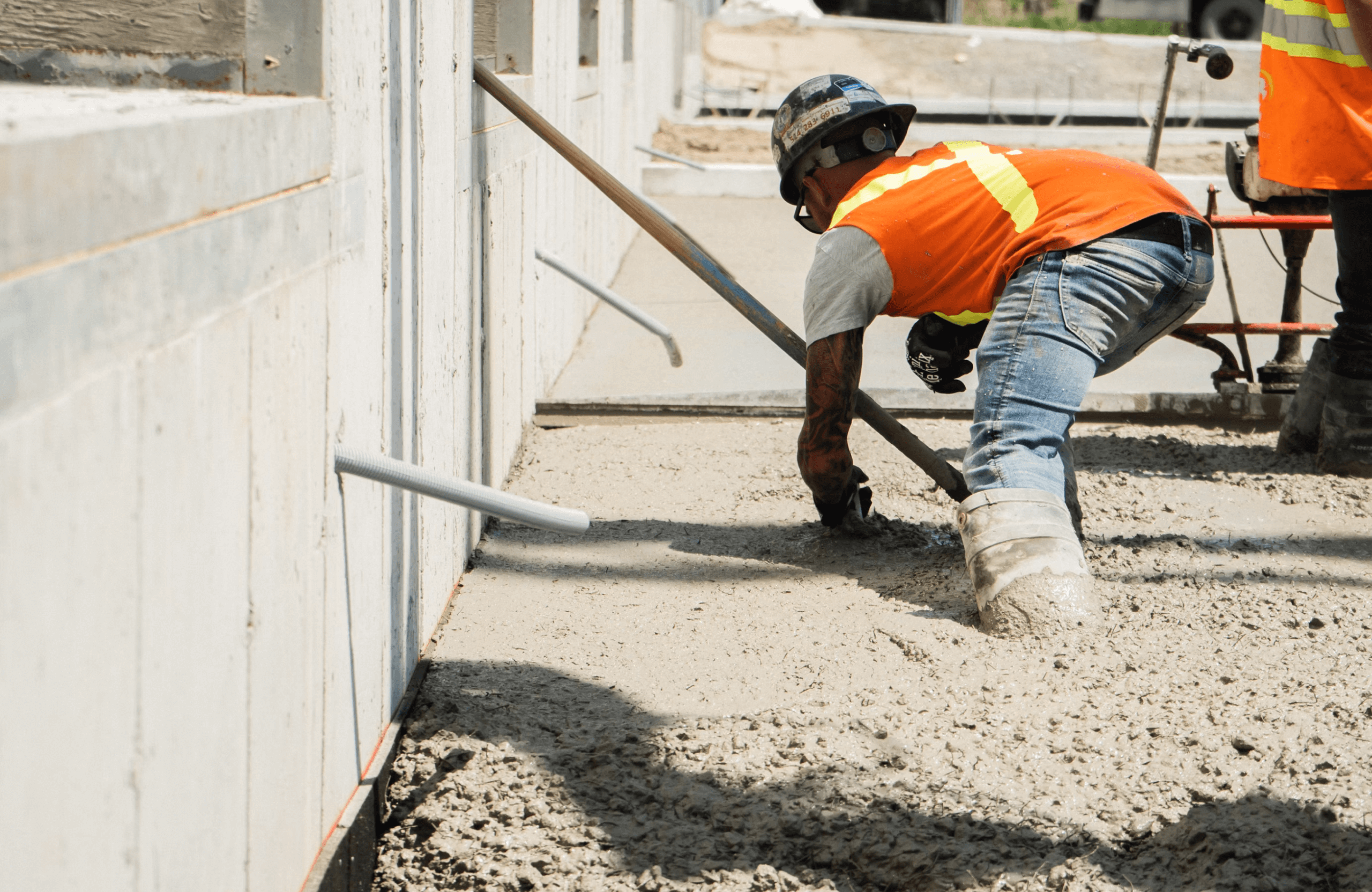 Stone Haven Developments Construction worker in an orange safety vest and helmet levels wet concrete with a long tool next to a building foundation. Ontario