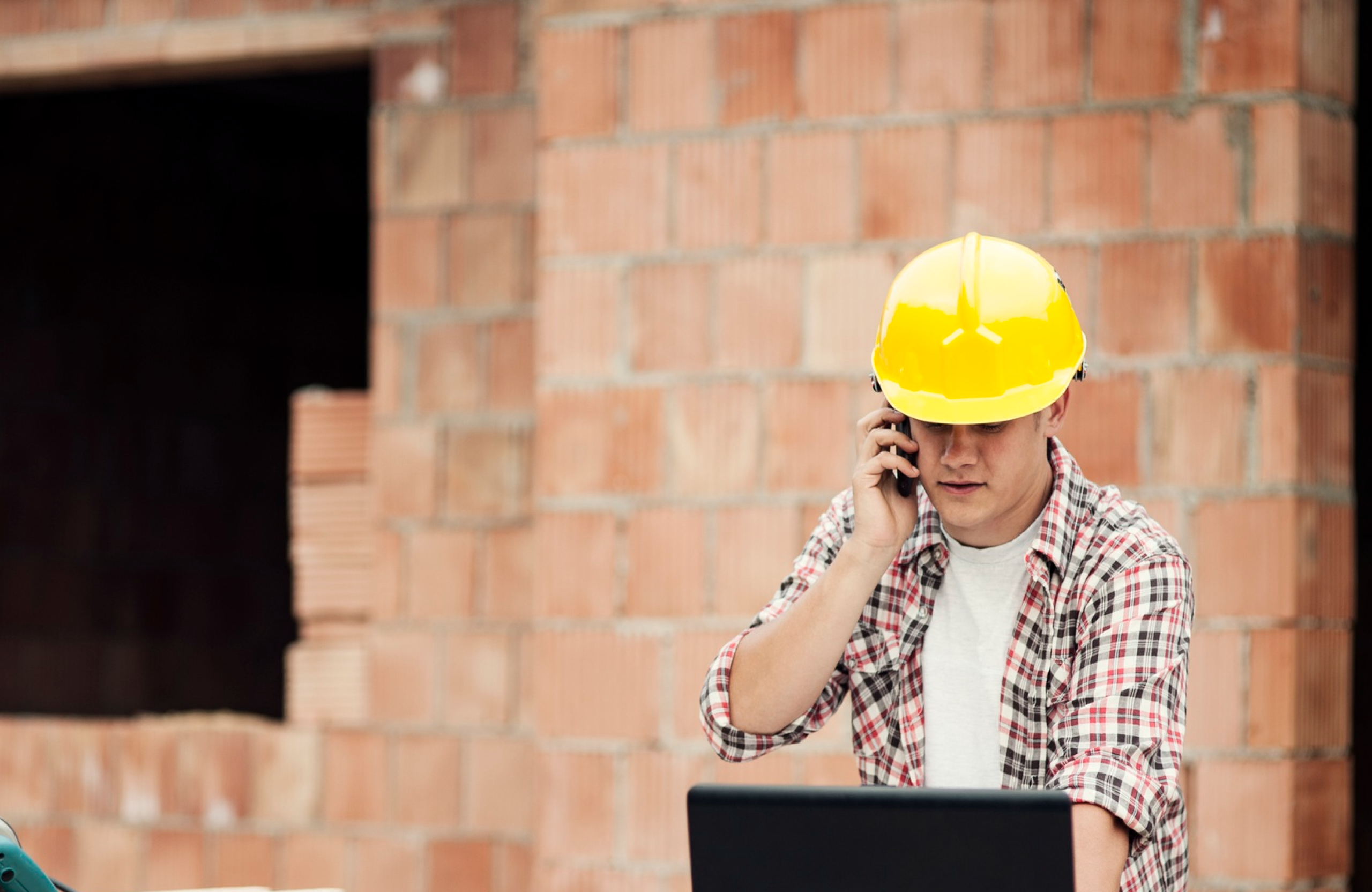 Stone Haven Developments A person wearing a yellow hard hat and plaid shirt talks on a phone while looking at a laptop, standing in front of an unfinished brick wall at a construction site. Ontario