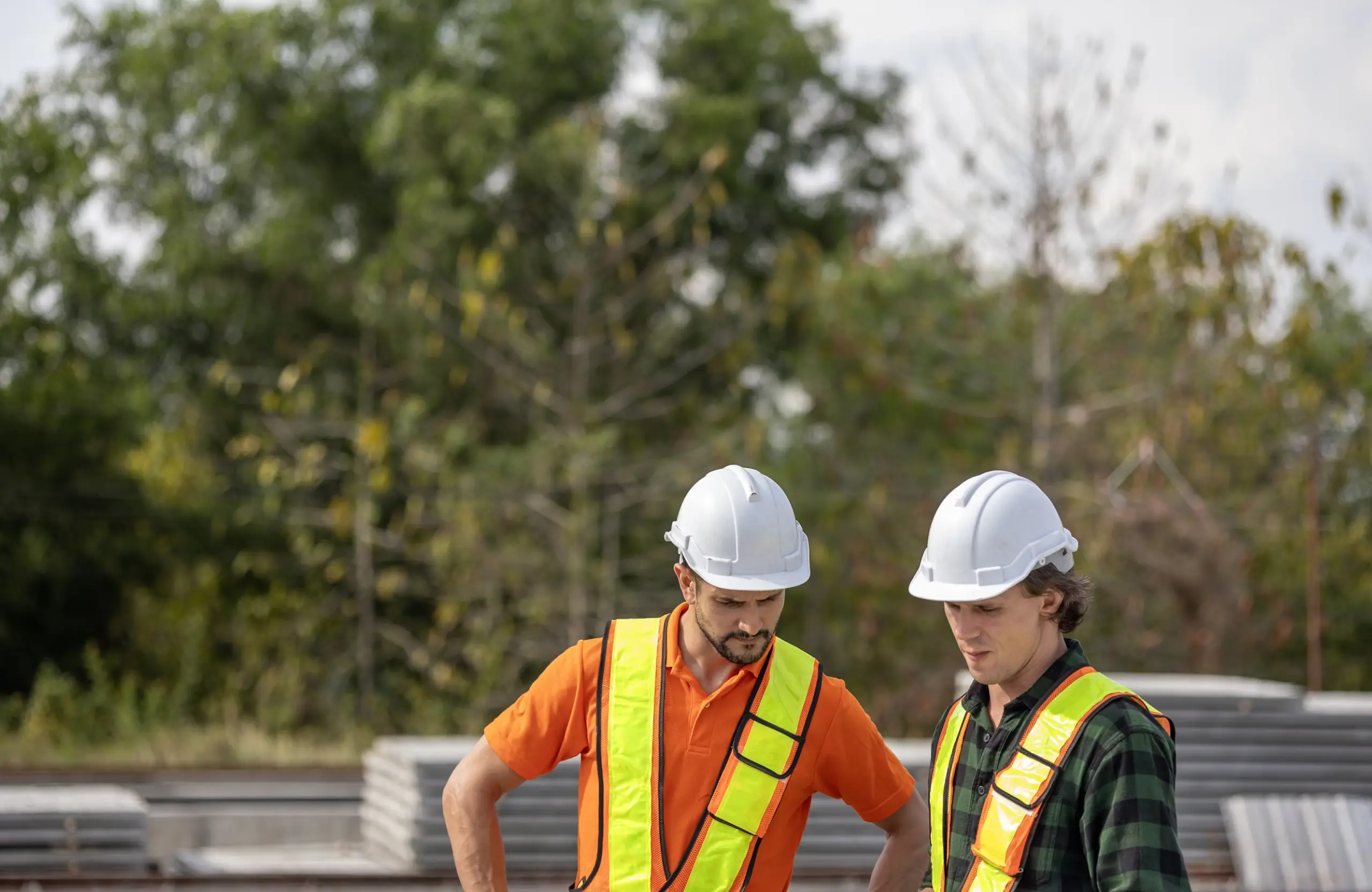 Stone Haven Developments Two construction workers wearing safety vests and helmets stand outdoors, looking down at something out of view, with trees and construction materials in the background. Ontario
