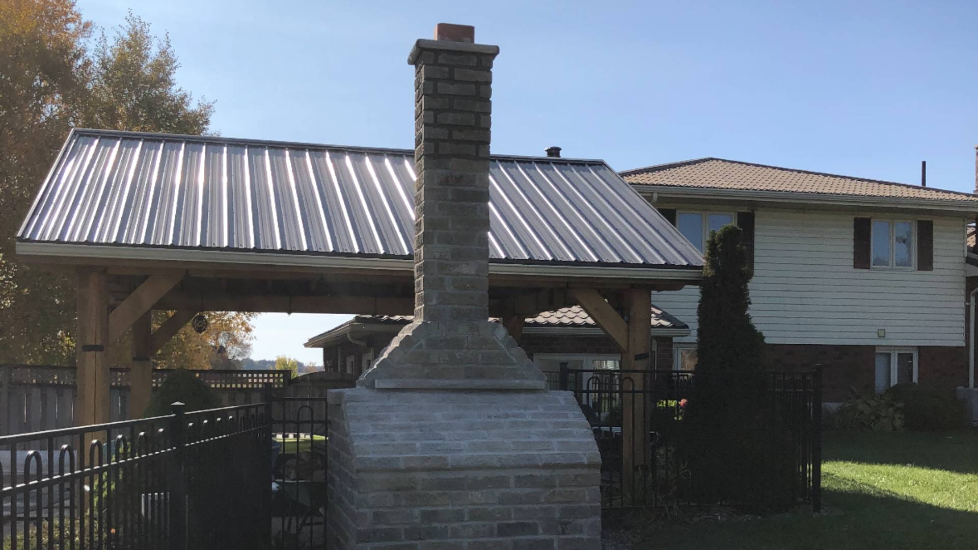 Stone Haven Developments A stone outdoor fireplace stands in front of a covered patio with a metal roof in a residential backyard. A house and trees are visible in the background. Ontario