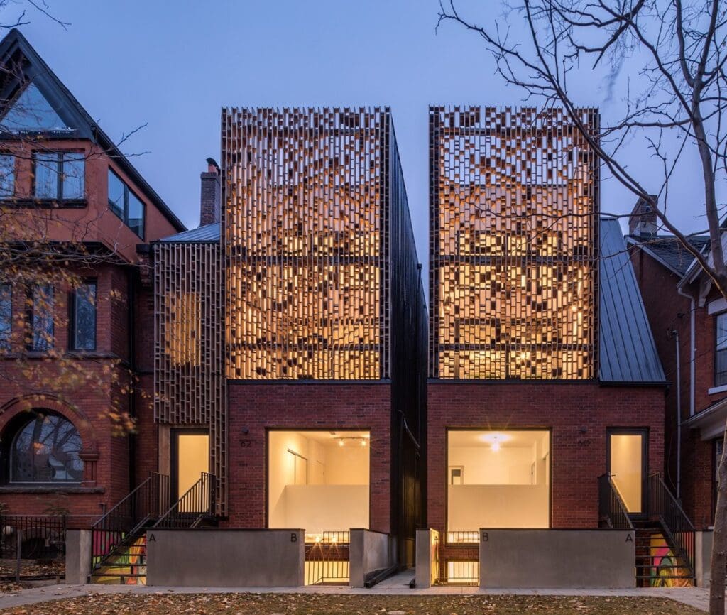 Stone Haven Developments Two modern townhouses with brick facades and decorative metal screens stand side by side between traditional-style homes at dusk. Ontario