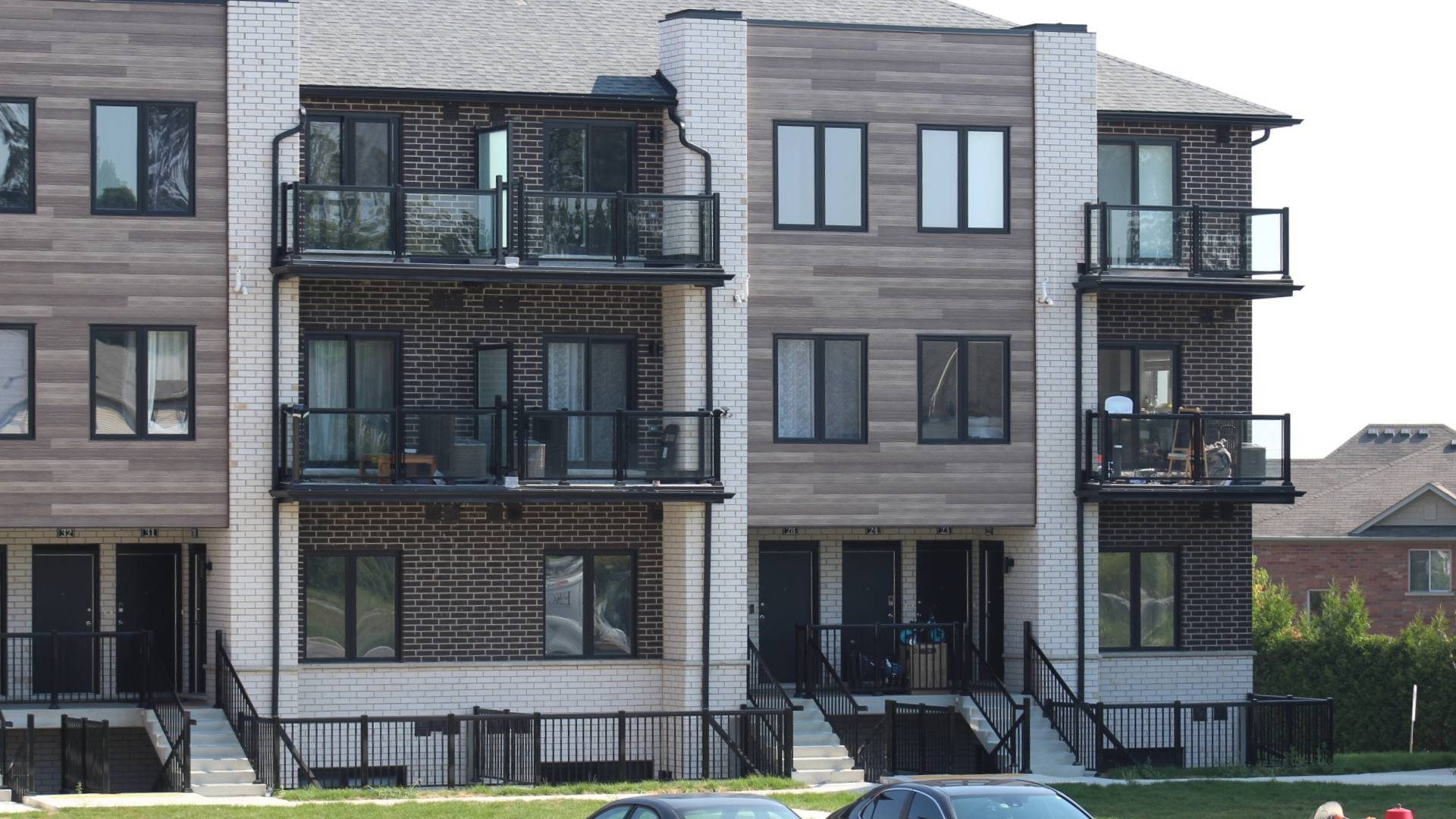 Stone Haven Developments A modern three-story apartment building with brick and wood panel exterior, several balconies, and black railings. Two cars are parked in front. Ontario
