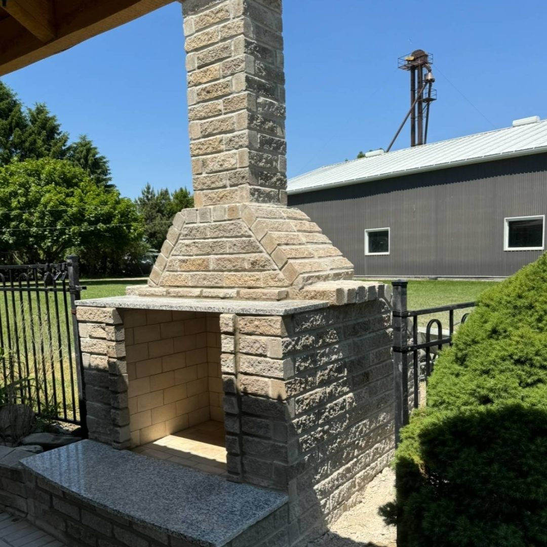 Stone Haven Developments Outdoor stone fireplace with a tall chimney and granite bench, situated on a patio near a metal fence, bushes, and a large building in the background. Ontario